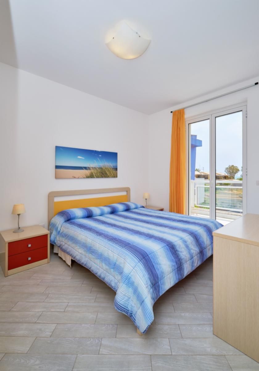 A bedroom with a bed, a nightstand, a desk, a window with curtains, and a painting on the wall.
