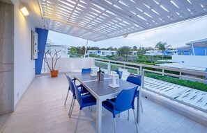 Uma área de refeições ao ar livre coberta, com mesa, cadeiras, um vaso de planta e vista para os edifícios próximos.