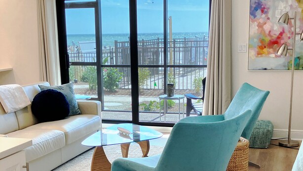 Ein modernes Wohnzimmer mit einem weißen Sofa, einem Glastisch und türkisfarbenen Sesseln. Ein großes Fenster bietet Blick auf den Ozean und einen umzäunten Bereich.