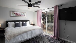 A bedroom with a bed, a ceiling fan, a large window with curtains, and a wall-mounted TV.