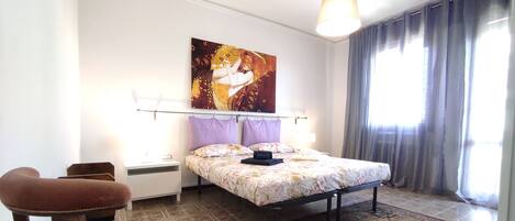 A bedroom with a bed, a chair, a nightstand, a lamp, a painting, and a window with curtains.