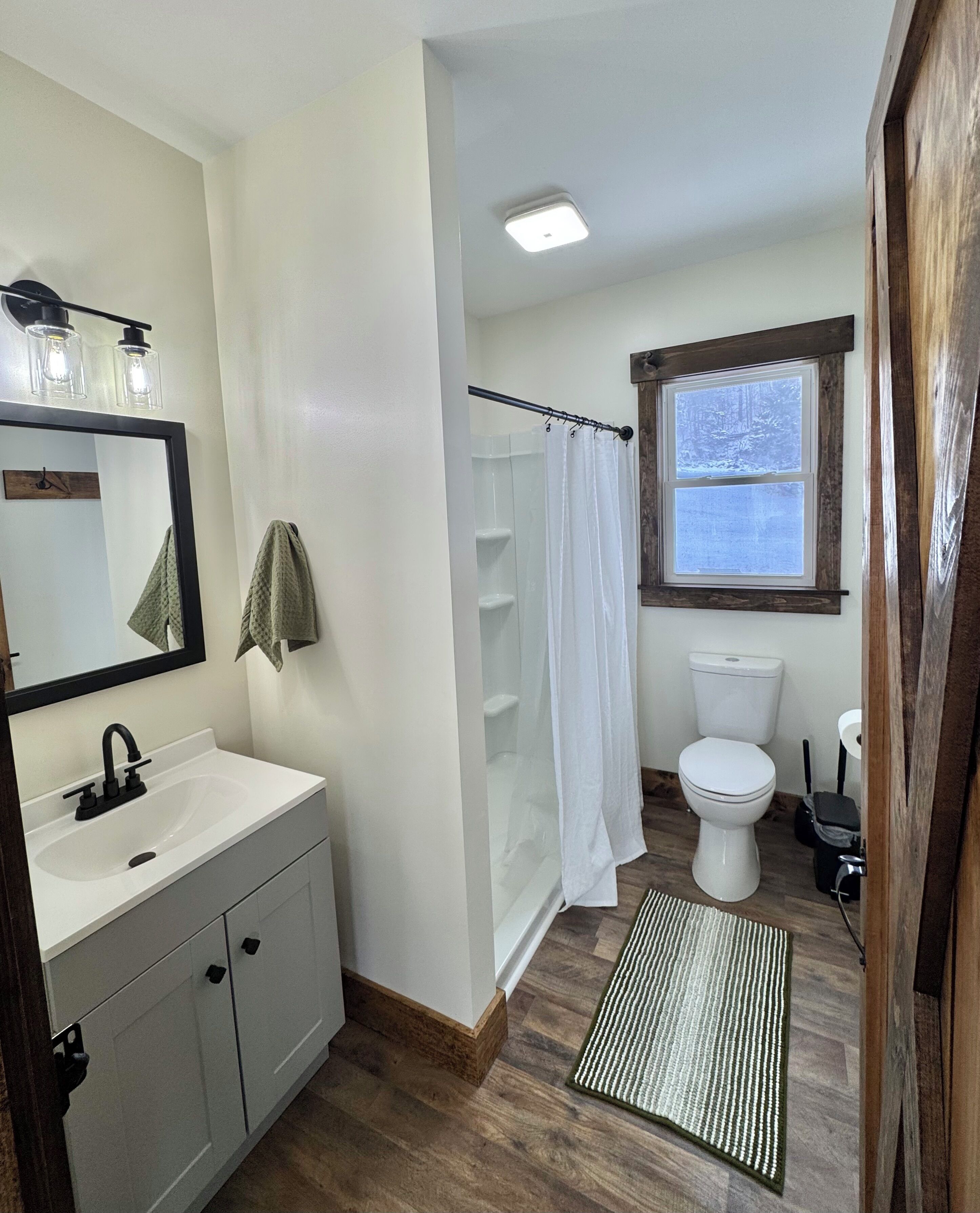 Une salle de bain avec un lavabo blanc, un miroir et des toilettes. On y trouve un rideau de douche et une fenêtre donnant sur des arbres.