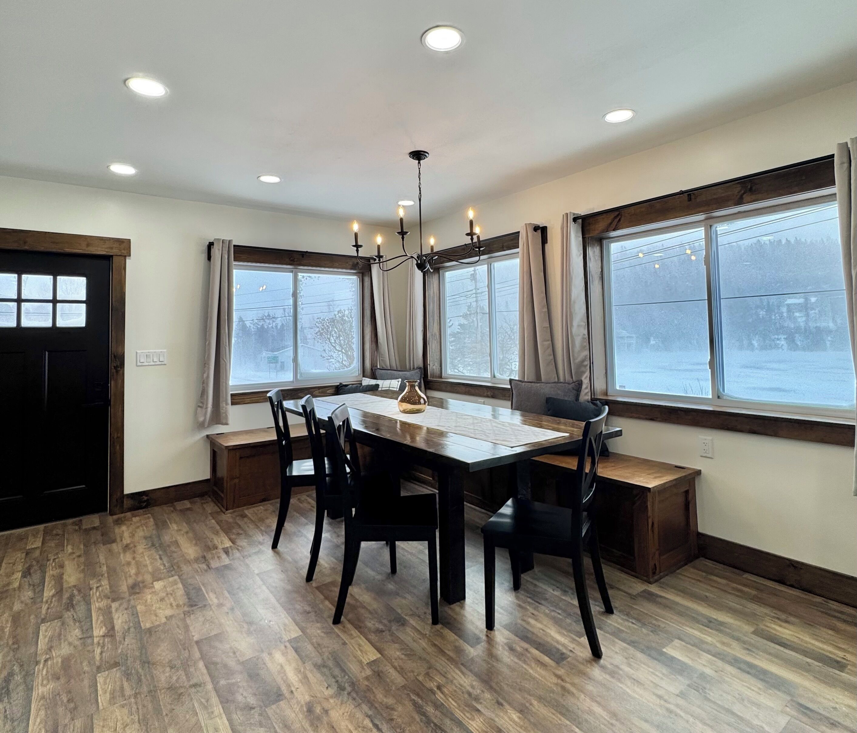 A dining room with a wooden table and chairs, large windows with curtains, and a chandelier.