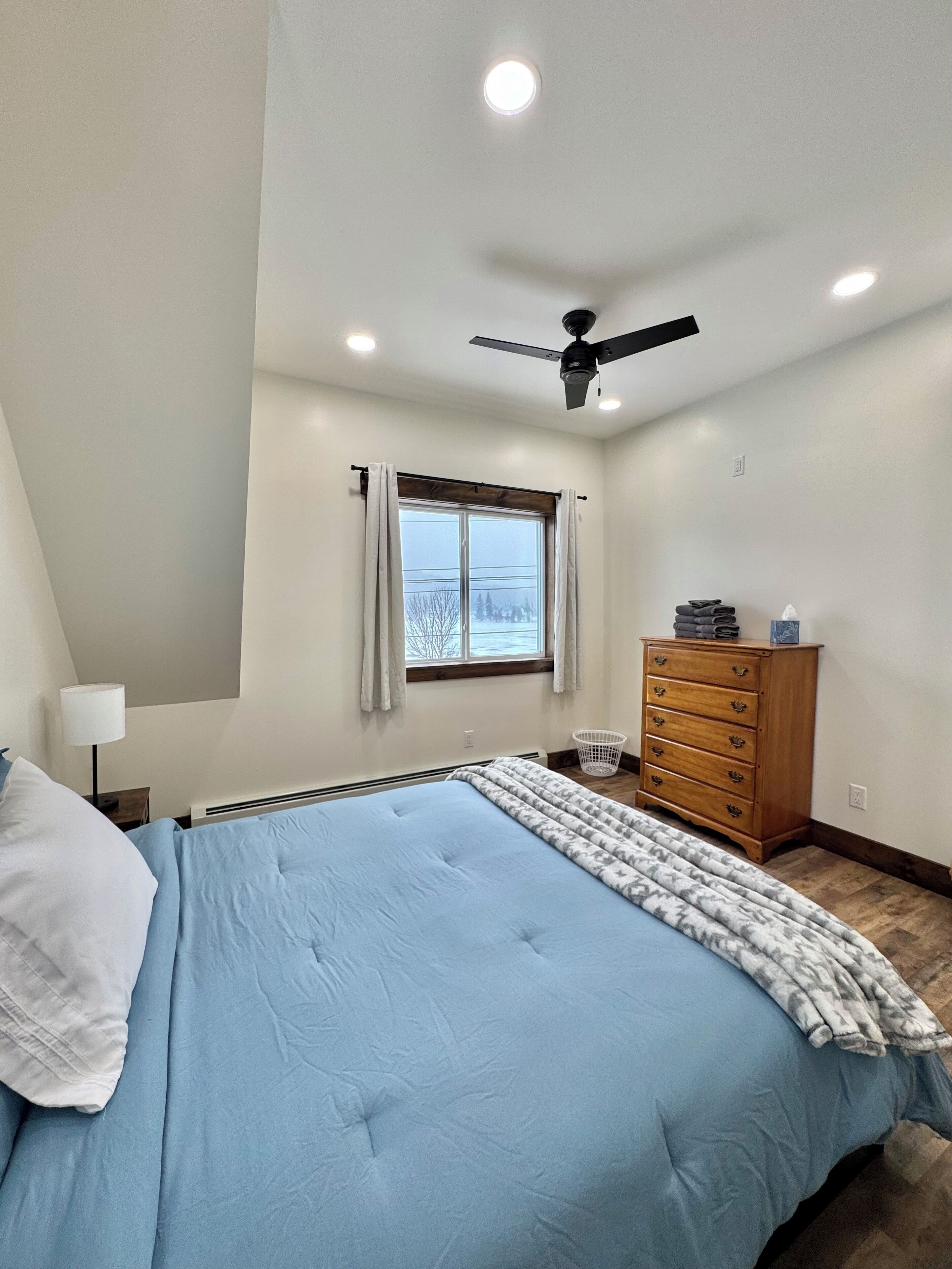 A bedroom with a bed, a dresser, a window with curtains, a ceiling fan, and recessed lighting.