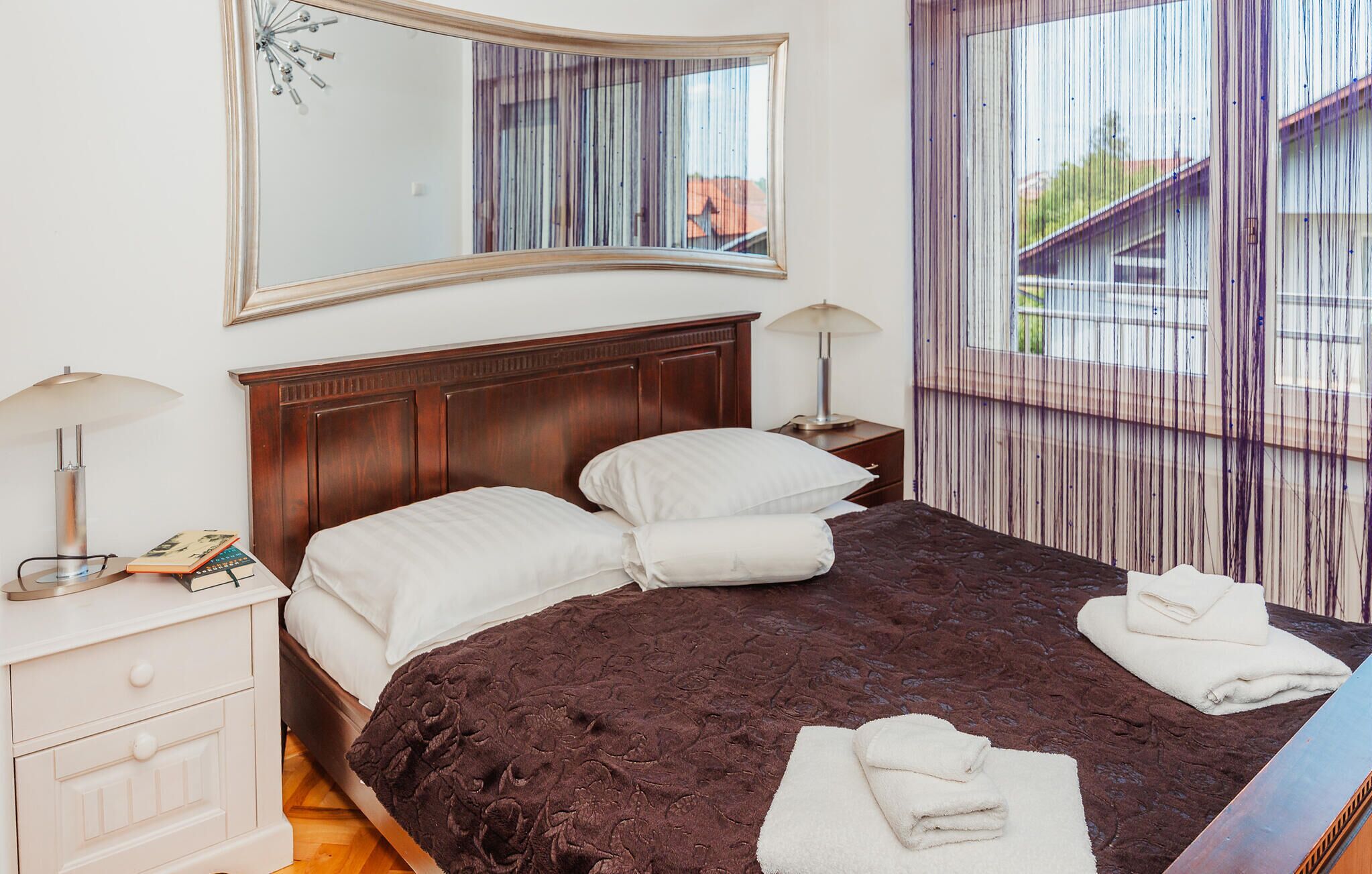 Een net opgemaakt bed met witte lakens, een donker beddekleed en twee witte handdoeken op het bed. Er staat een houten nachtkastje met een lamp en een boek naast het bed. Een grote spiegel hangt boven het bed en weerspiegelt de kamer.