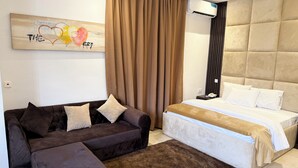 Un dormitorio moderno con un sofá marrón, un reposapiés a juego, una cama grande con ropa de cama blanca y un aire acondicionado de pared.