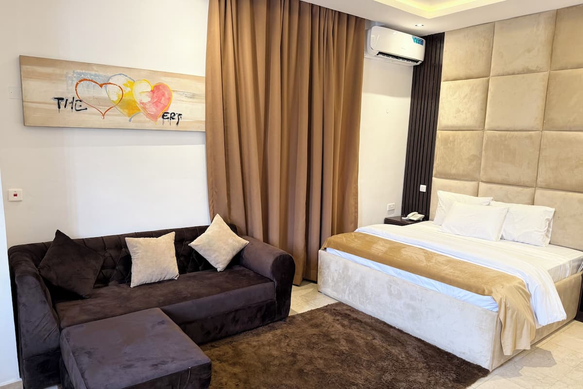 Une chambre moderne avec un canapé marron, un pouf assorti, un grand lit avec une literie blanche et un climatiseur fixé au mur.