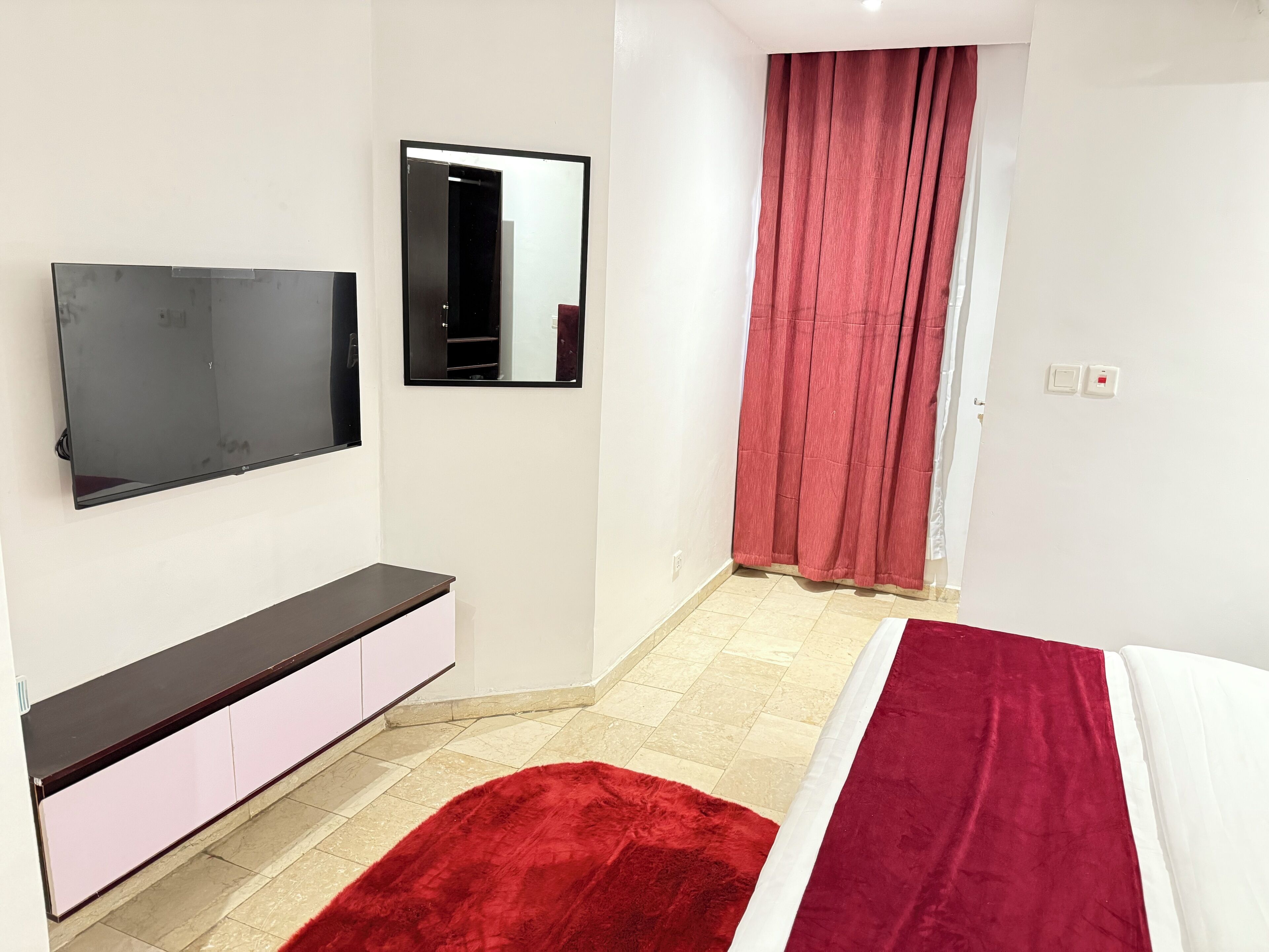 Une chambre à coucher avec un lit, une télévision, un miroir, un rideau rouge et un mur blanc.