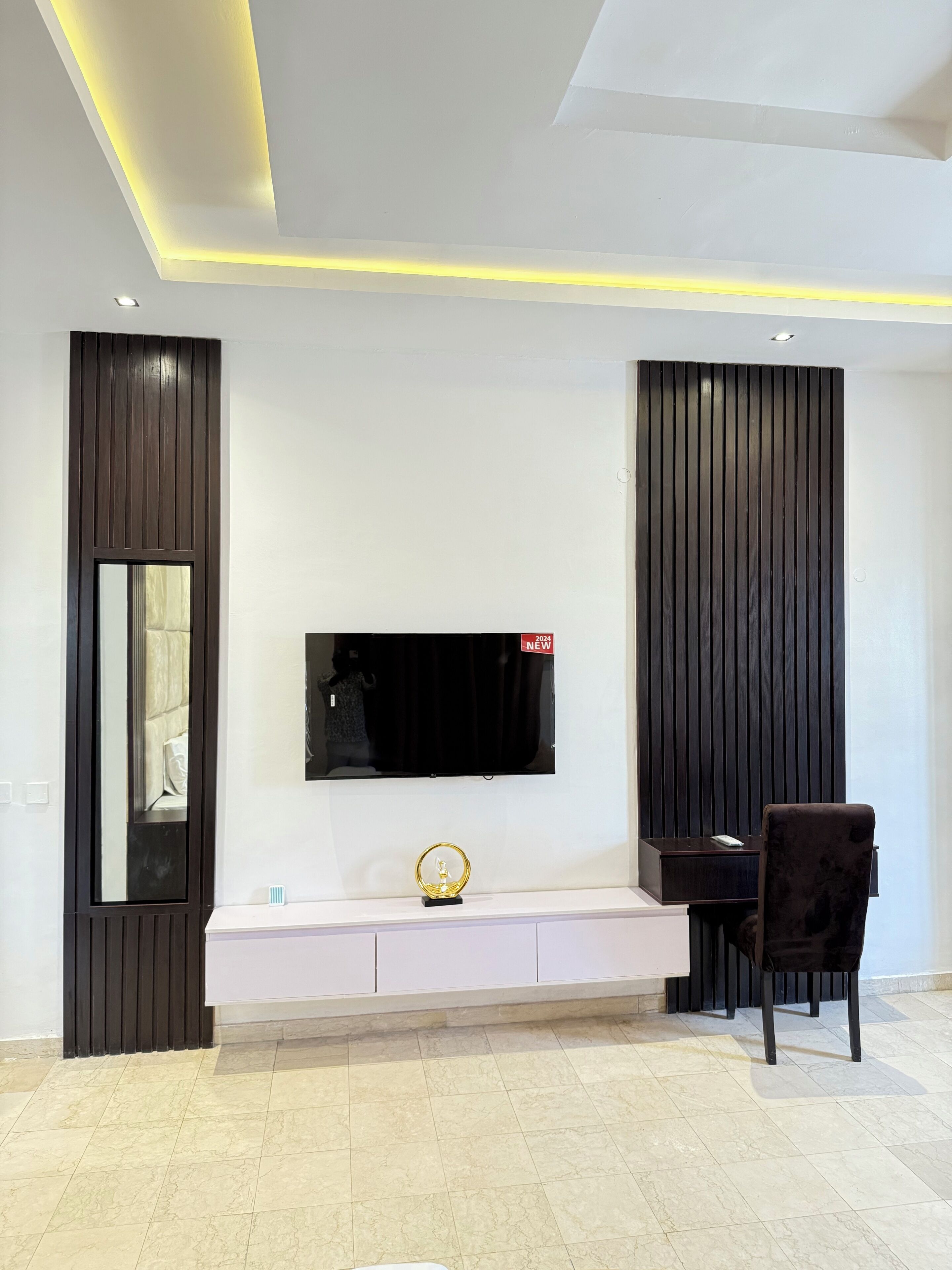 A modern living room with a flat-screen TV mounted on a white wall, a dark wooden accent wall, a large mirror, a white console table with a decorative sphere, and a dark upholstered chair.