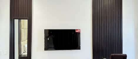 A modern living room with a flat-screen TV mounted on a white wall, a dark wooden accent wall, a large mirror, a white console table with a decorative sphere, and a dark upholstered chair.