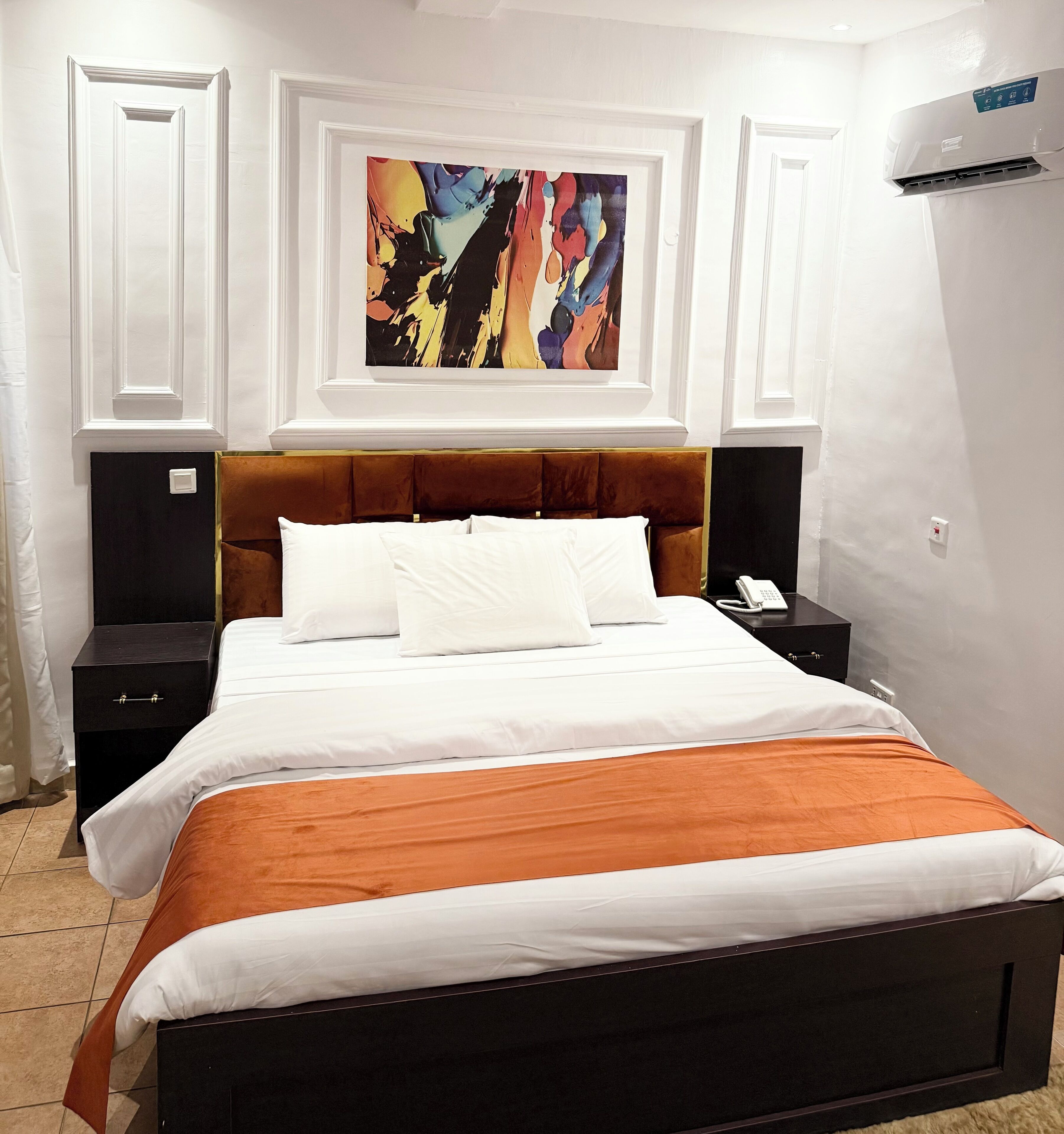 A hotel room with a large bed, white bedding, a brown headboard, a colorful abstract painting above the bed, and a white air conditioning unit on the wall.