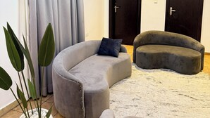 Una sala de estar moderna con un sofá gris curvo, un reposapiés gris más pequeño y una planta en maceta. Hay dos puertas de madera oscura y una lámpara de techo con iluminación LED azul.