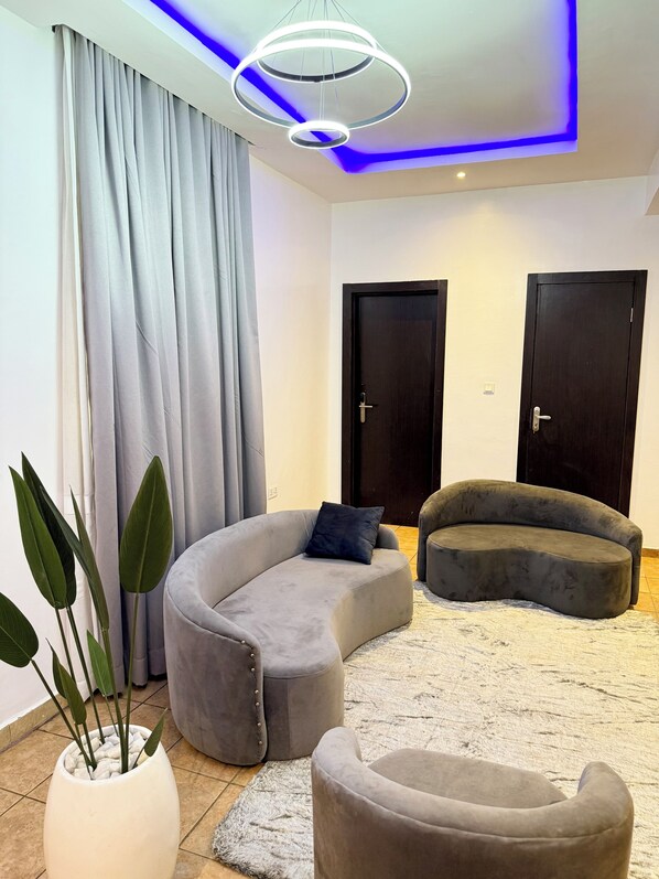 Una sala de estar moderna con un sofá gris curvo, un reposapiés gris más pequeño y una planta en maceta. Hay dos puertas de madera oscura y una lámpara de techo con iluminación LED azul.