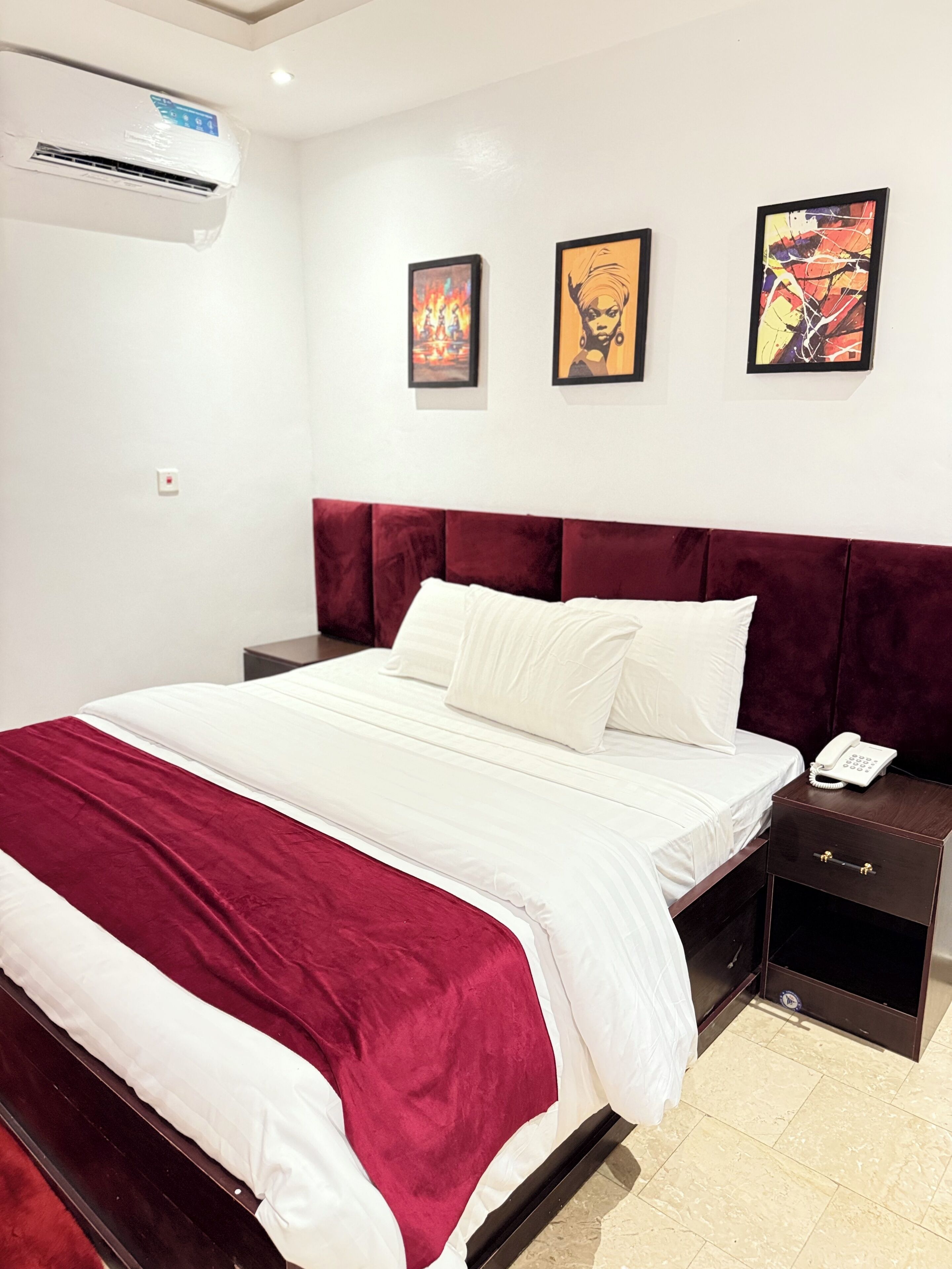 A hotel room with a large bed, a red headboard, a bedside table with a phone, and three framed artworks on the wall.
