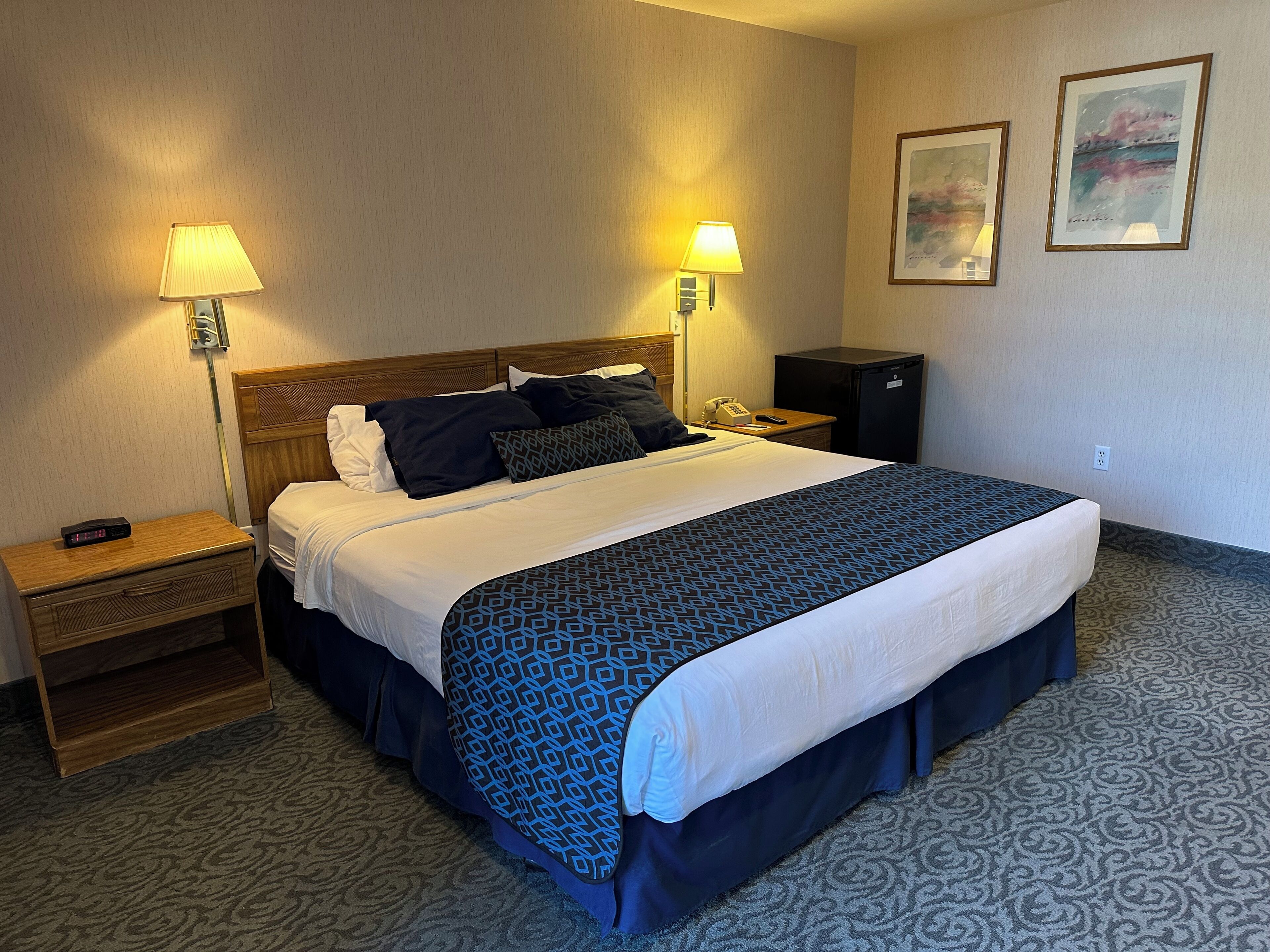 Une chambre d’hôtel avec un grand lit, deux lampes de chevet, une table de chevet, un téléphone, un petit réfrigérateur et deux œuvres d’art encadrées accrochées au mur.