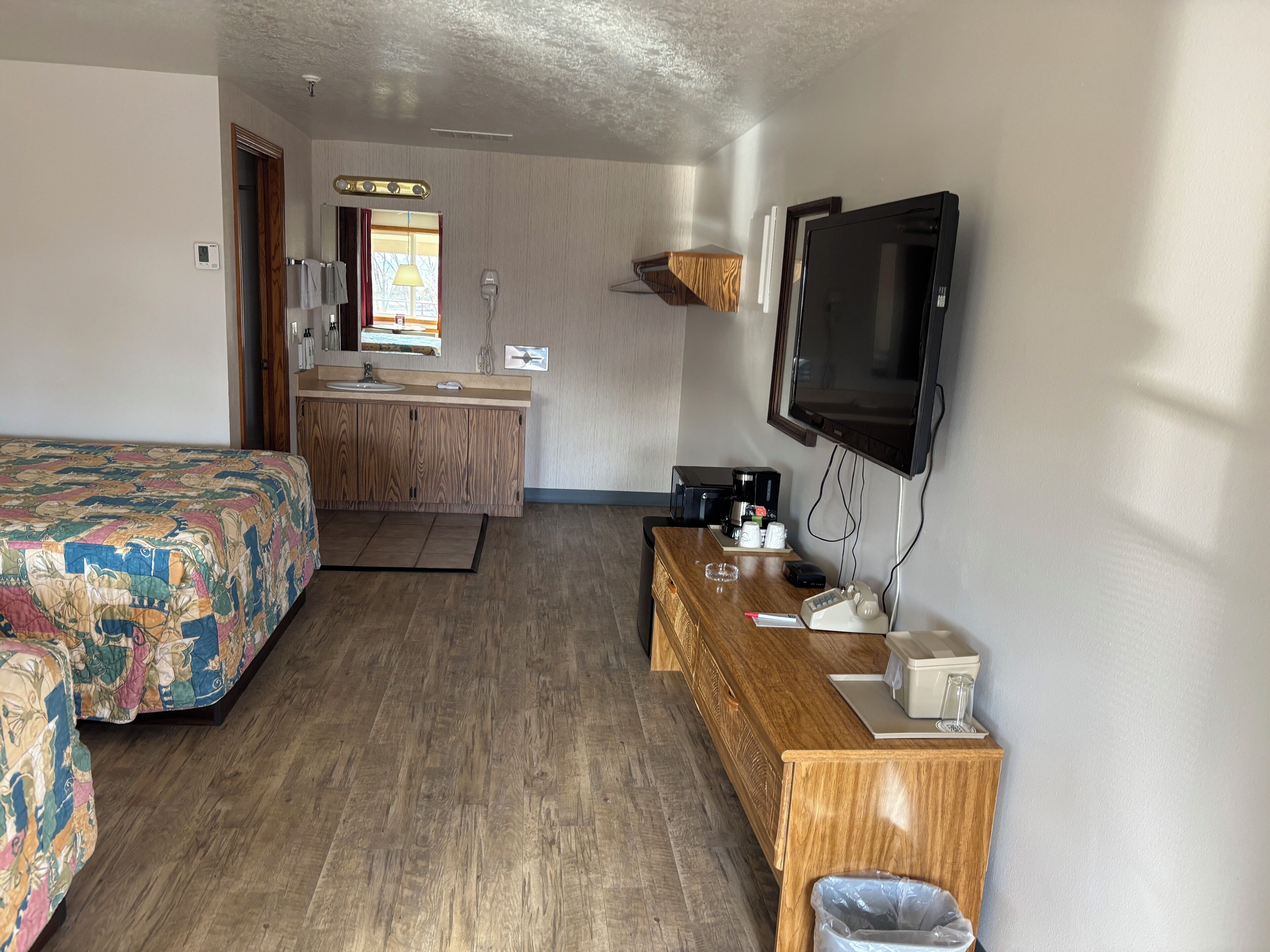 A hotel room with a bed, a wooden desk, a television, a coffee maker, and a bathroom visible in the background.