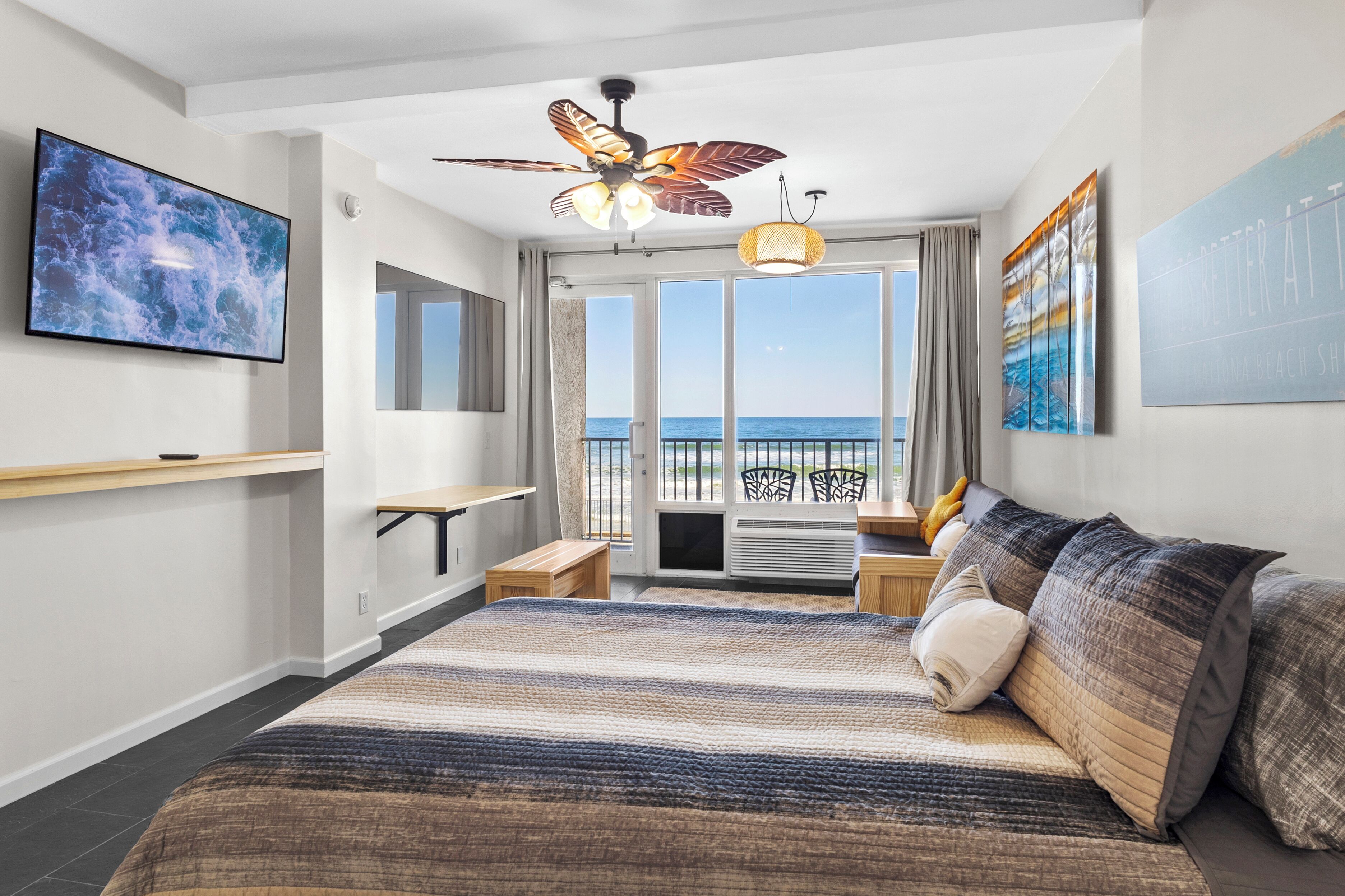 A modern hotel room with a large bed, a ceiling fan, a flat-screen TV, a desk with a chair, and a view of the ocean.