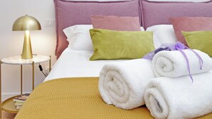 Una cama bien hecha con toallas blancas dobladas, un plaid color mostaza y una lámpara dorada sobre una mesita.