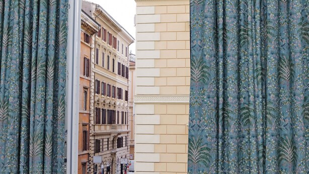 Vista a través de una ventana con cortinas estampadas, mostrando un edificio de varios pisos y balcones.