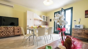 Cocina moderna con armarios blancos, comedor con sillas blancas, una mesa de comedor de madera y un televisor sobre un mueble de madera.