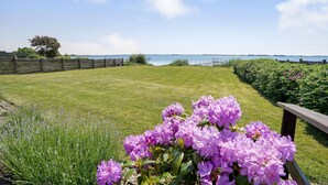 A grassy lawn with a wooden fence, a flower bed with purple flowers, and a view of the sea. - Gorgeous home in Faaborg with sauna (Faaborg)