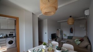 A modern kitchen with a washing machine, a dining area with a table set for two, and a living area with a sofa and a TV.