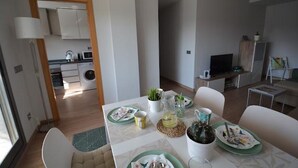 A modern kitchen with white cabinets, a microwave, and a sink. There is a dining area with a table set for two, featuring green plates and a vase with flowers.