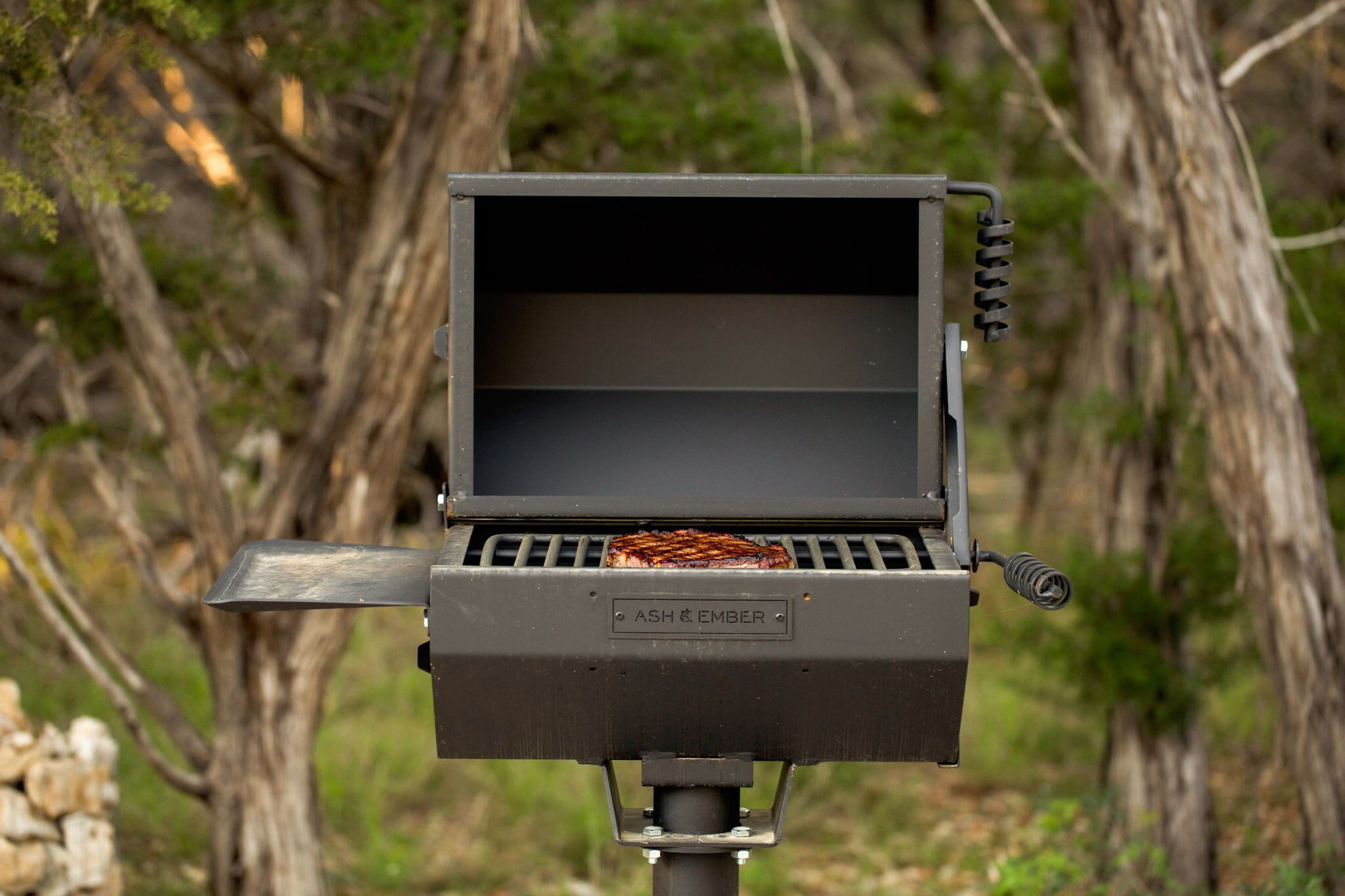 A portable grill with a single steak cooking, surrounded by trees.