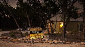 Placa com a inscrição 'STILLWATER' em um cenário natural, com árvores e um edifício ao fundo.