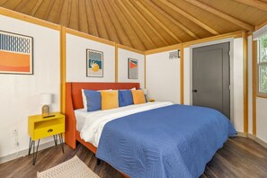 A bedroom with a wooden ceiling, a bed with blue and orange bedding, a yellow nightstand, and framed artwork on the walls.
