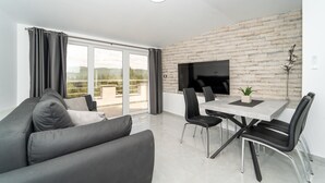 Un salon moderne avec un canapé gris, une table à manger avec quatre chaises, un téléviseur à écran plat fixé à un mur dont le motif rappelle celui d’un mur de briques, et un balcon avec des rideaux.