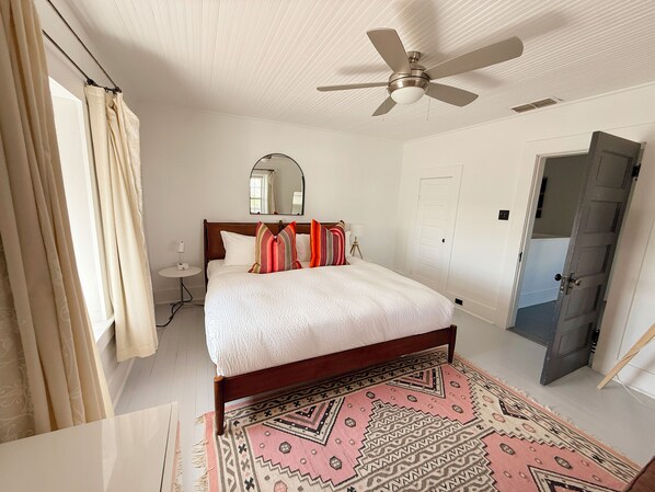 A bedroom with a bed, a ceiling fan, a bedside table, a mirror, a door, and a patterned rug.