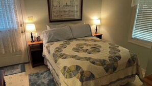 Een keurig opgemaakt bed met een sprei, twee bedlampen, een lijst met een afbeelding aan de muur en een raam met jaloezieën.
