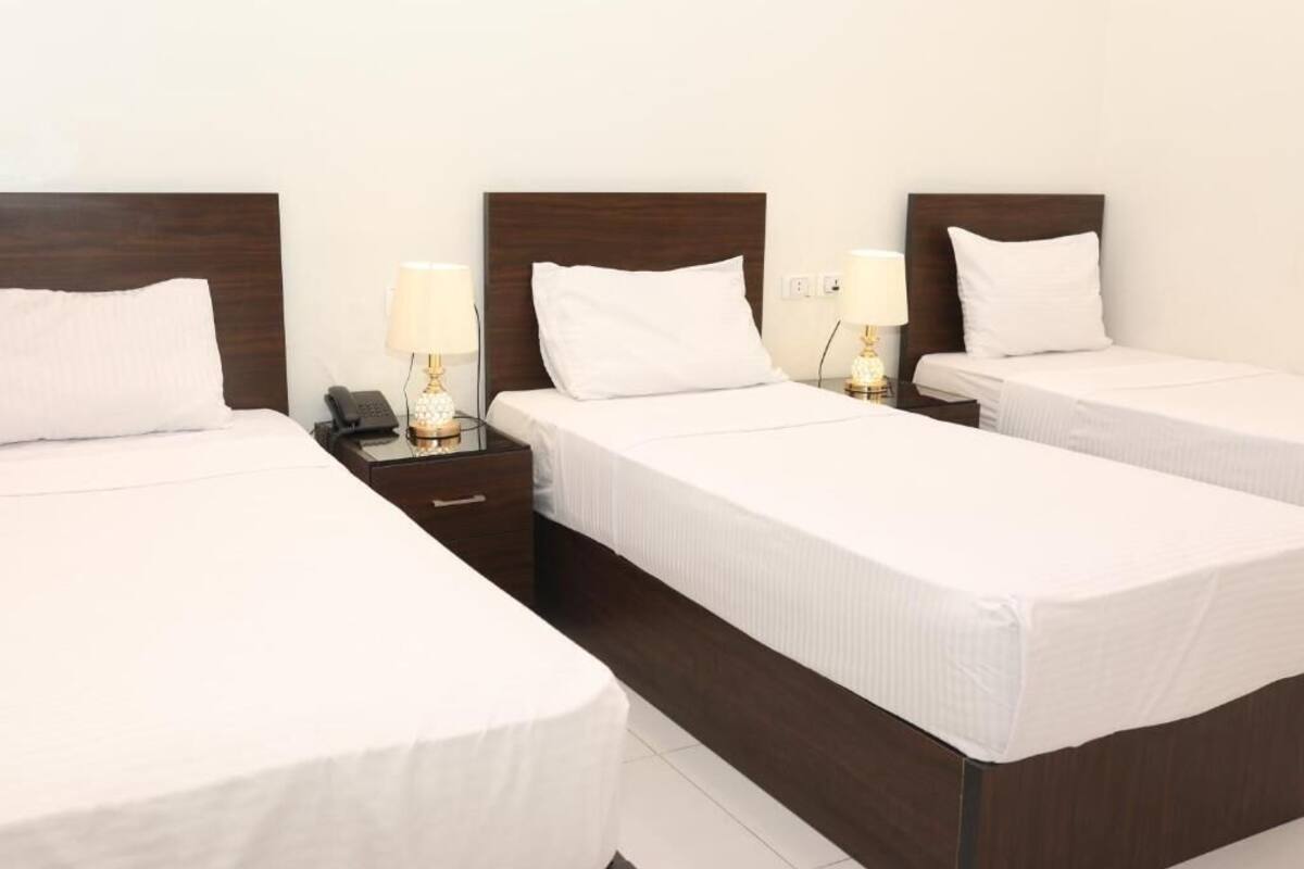 Ein Hotelzimmer mit drei Einzelbetten, jedes mit einem Holz-Kopfschild und weißer Bettwäsche. An der linken Seite befinden sich Nachttische mit Lampen und einem Telefon.