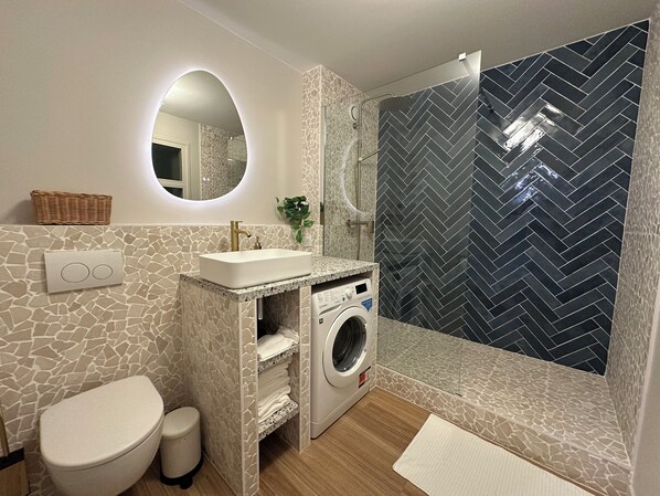 Une salle de bain moderne avec un mur imitant la pierre, un lave-linge encastré, un lavabo avec un robinet doré et une douche dont le carrelage est disposé en motif chevrons.