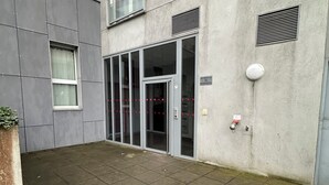 Modernilla designilla varustettu rakennus, jossa on lasinen sisäänkäyntiovi, pieni ulkoalue laatoitetulla lattialla ja ruukussa oleva kasvi.