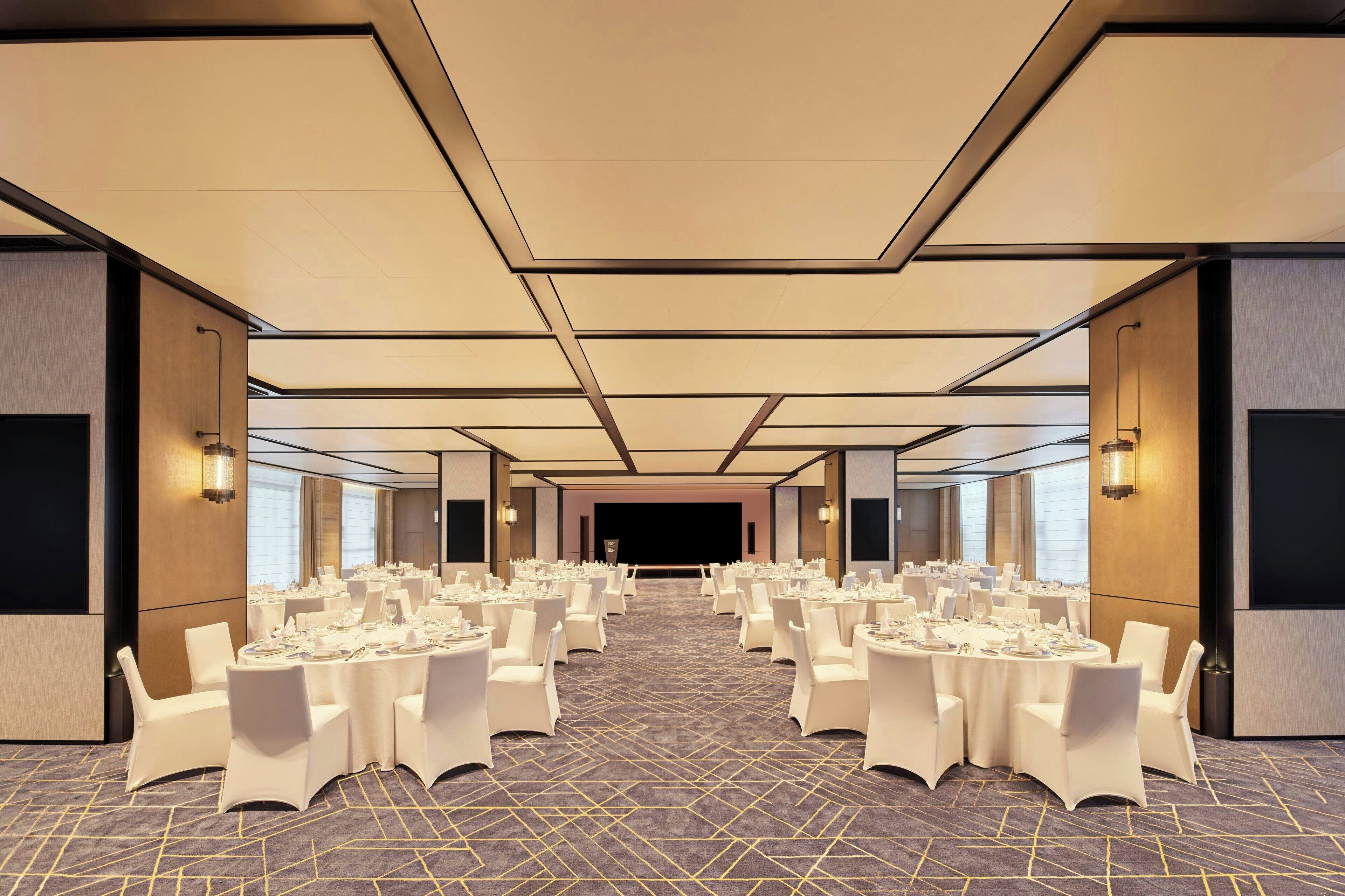 A large, well-lit conference room with round tables and chairs, a stage at the front, and a carpeted floor.