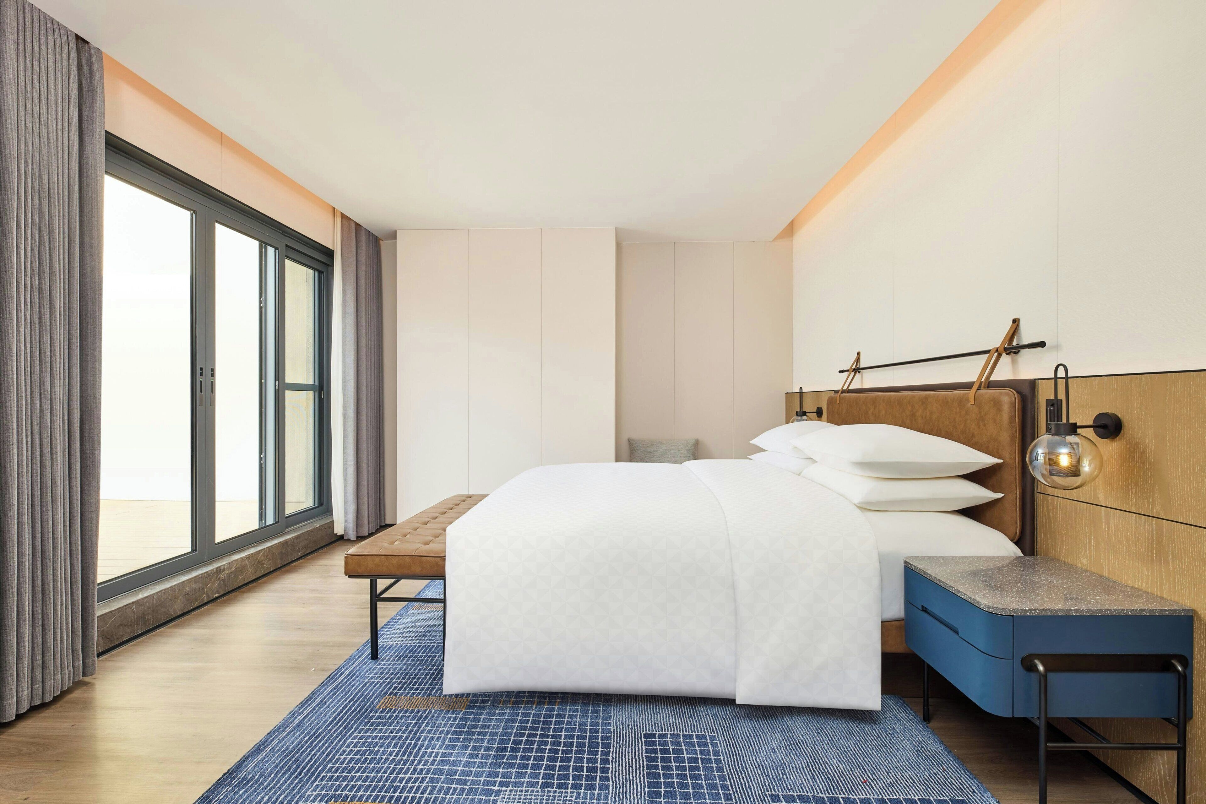 A modern hotel room with a large bed, a wooden headboard, a nightstand, and a large window offering a view.