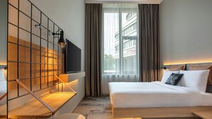 A modern hotel room with a large bed, a TV mounted on a grid wall, a small round table, and a window with curtains. - Moxy Hamburg - Finkenwerder (Hamburg)