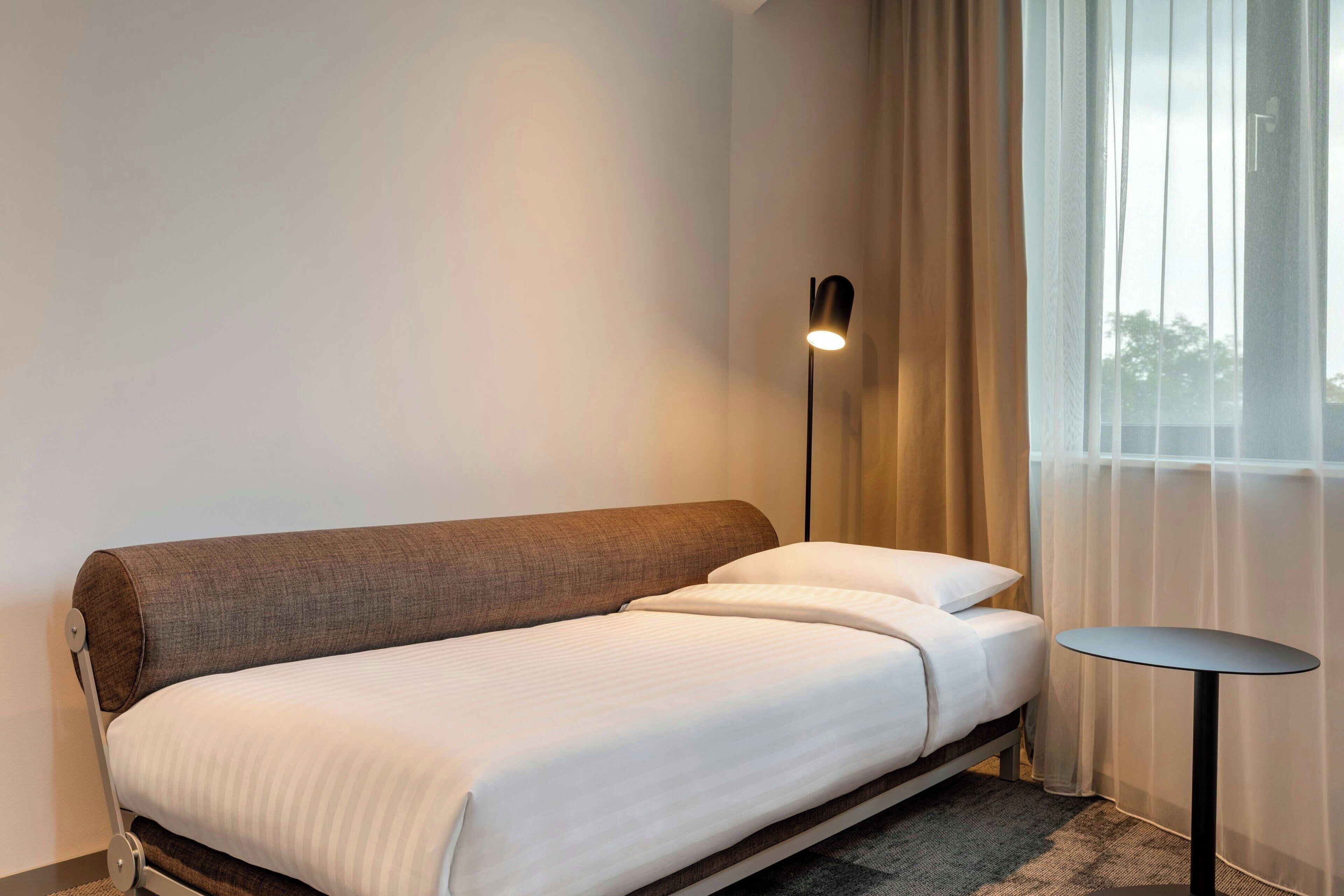 Ein modernes Hotelzimmer mit einem Einzelbett, einem kleinen runden Tisch und einer Stehlampe.
