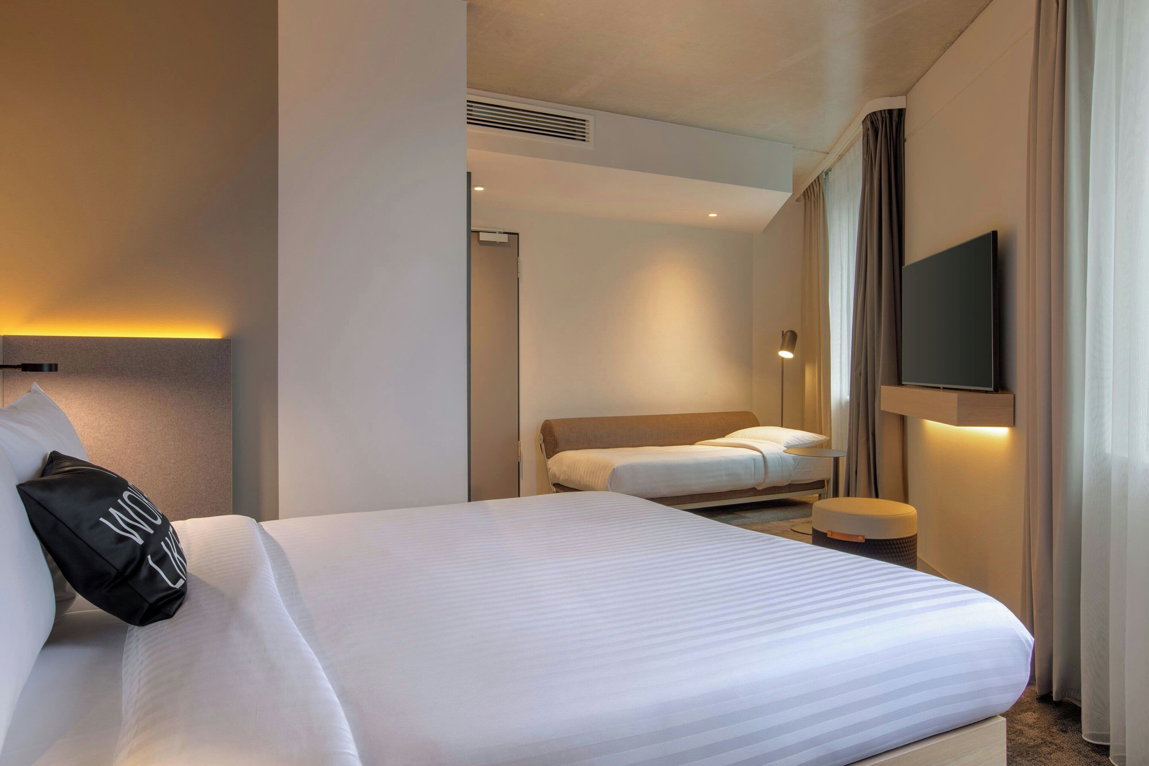 A hotel room with a large bed, a bedside table, a TV, and a wall-mounted light.