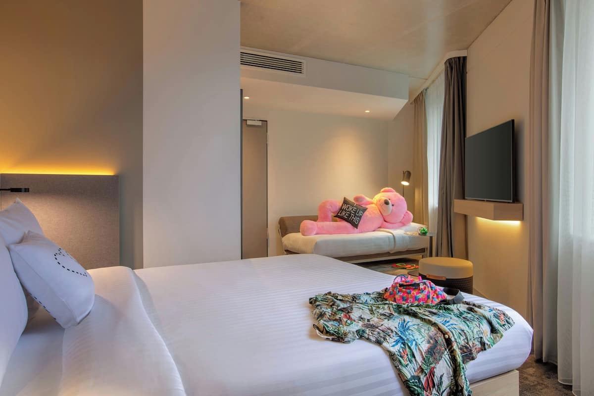 Una habitación de hotel con una cama grande, una mesita de noche con lámpara, un televisor montado en la pared y un oso de peluche rosa suave sobre la cama.