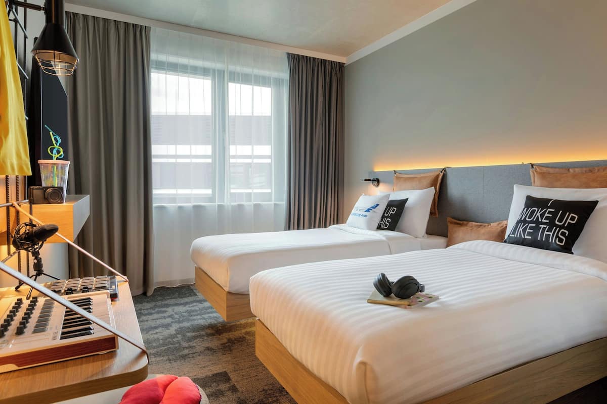 Habitación de hotel con dos camas, un escritorio con teclado, una lámpara y vistas al exterior a través de amplias ventanas.