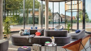 A modern living room with a curved ceiling, large windows, and a view of a street and buildings. - Moxy Hamburg - Finkenwerder (Hamburg)