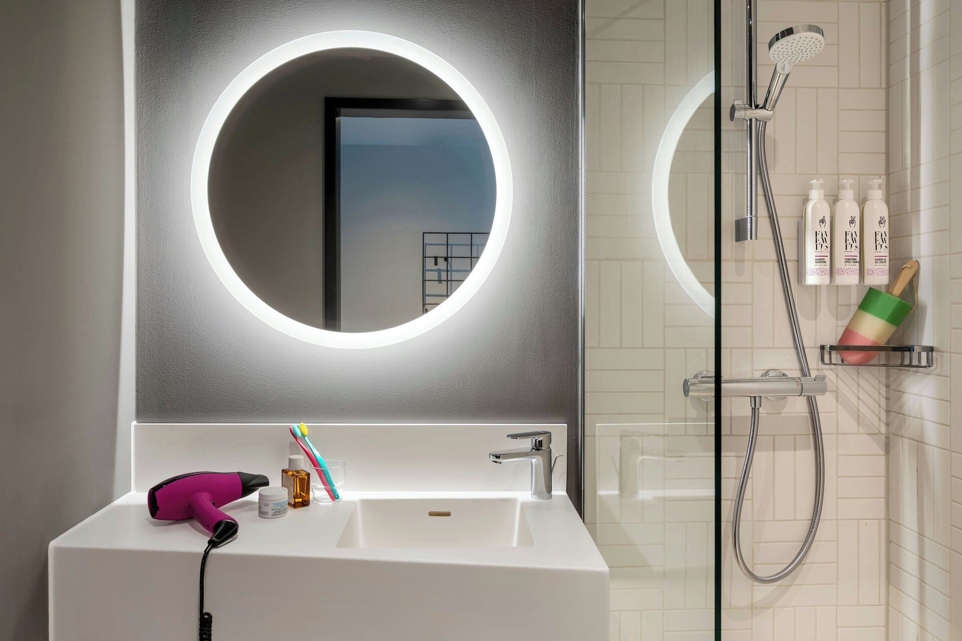 A bathroom with a white sink, a mirror with a circular light, a handheld showerhead, and toiletries on a shelf.