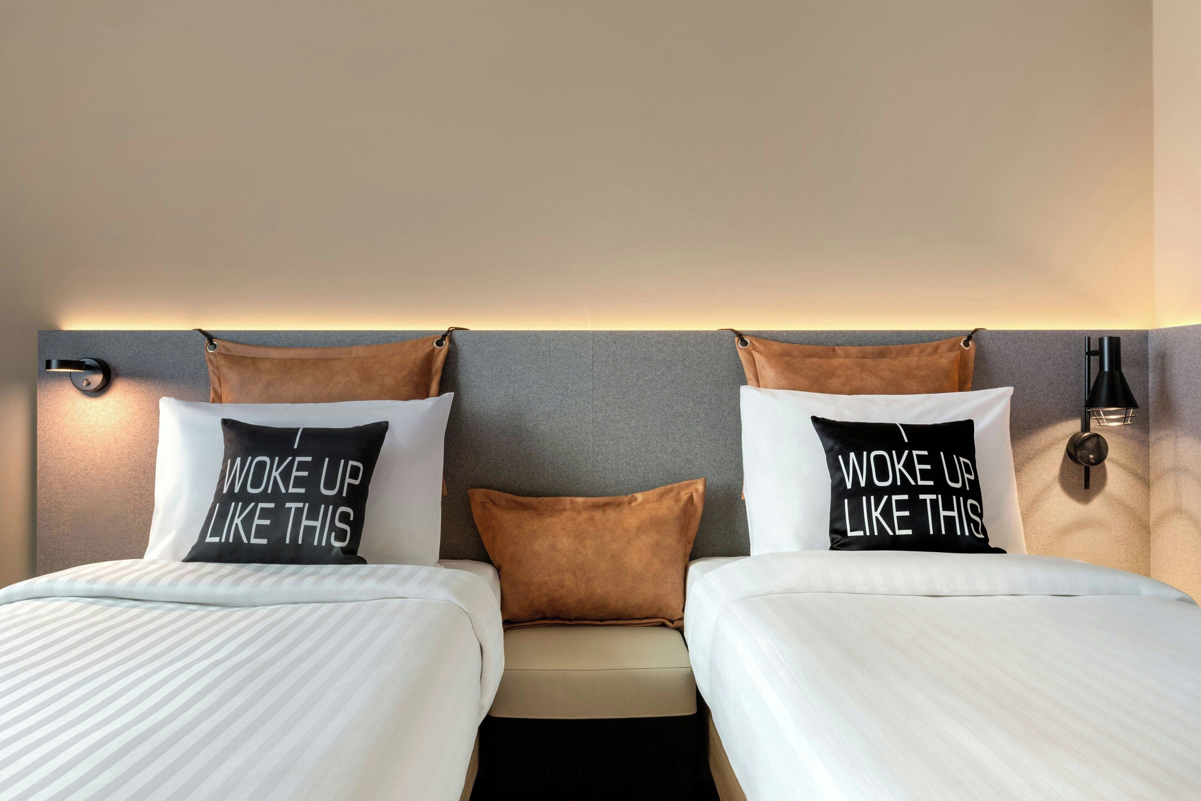 A hotel room with two beds, each with a pillow that reads 'Woke up like this.'