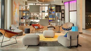 A modern living room with contemporary furniture, a large bookshelf, and a vibrant light installation. - Moxy Hamburg - Finkenwerder (Hamburg)