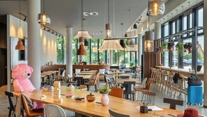 A spacious cafe with multiple tables and chairs, a large teddy bear decoration, hanging pendant lights, and large windows offering a view of the outdoors. - Moxy Hamburg - Finkenwerder (Hamburg)