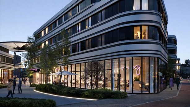 A modern multi-story building with a curved facade, large glass windows, and a well-lit entrance area with outdoor seating. - Moxy Hamburg - Finkenwerder (Hamburg)