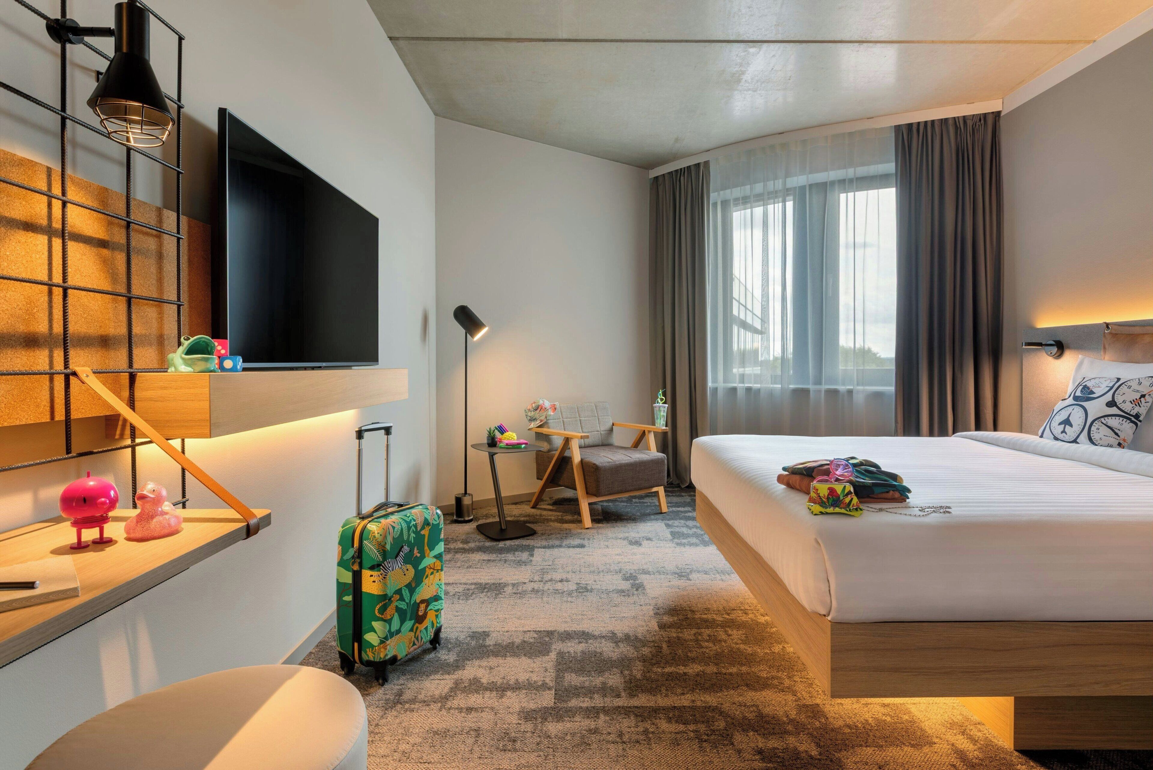 Habitación de hotel con cama, escritorio, silla, televisor y maleta.